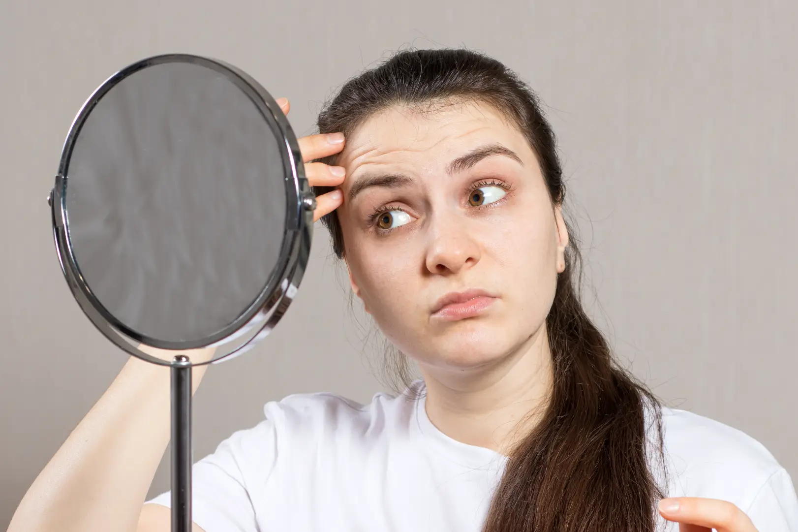 Woman looks toward a round mirror while touching visible horizontal lines on her forehead.