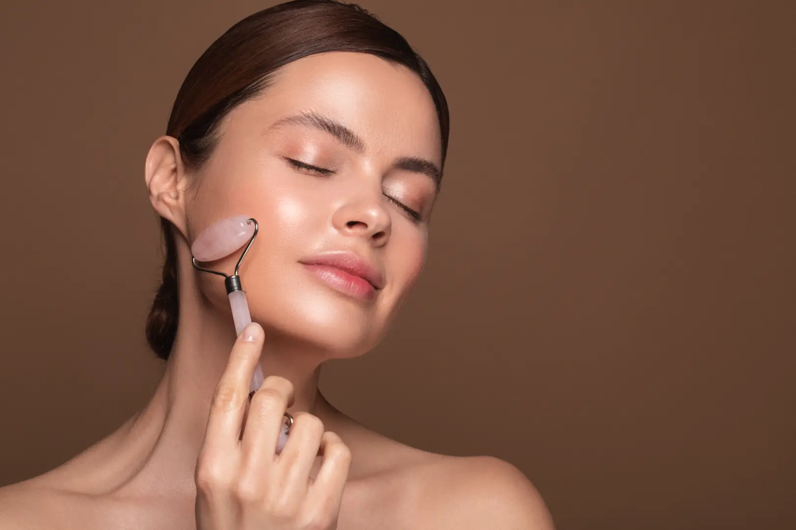 Woman with closed eyes gently rolls a pink facial roller along her cheek against a brown background.
