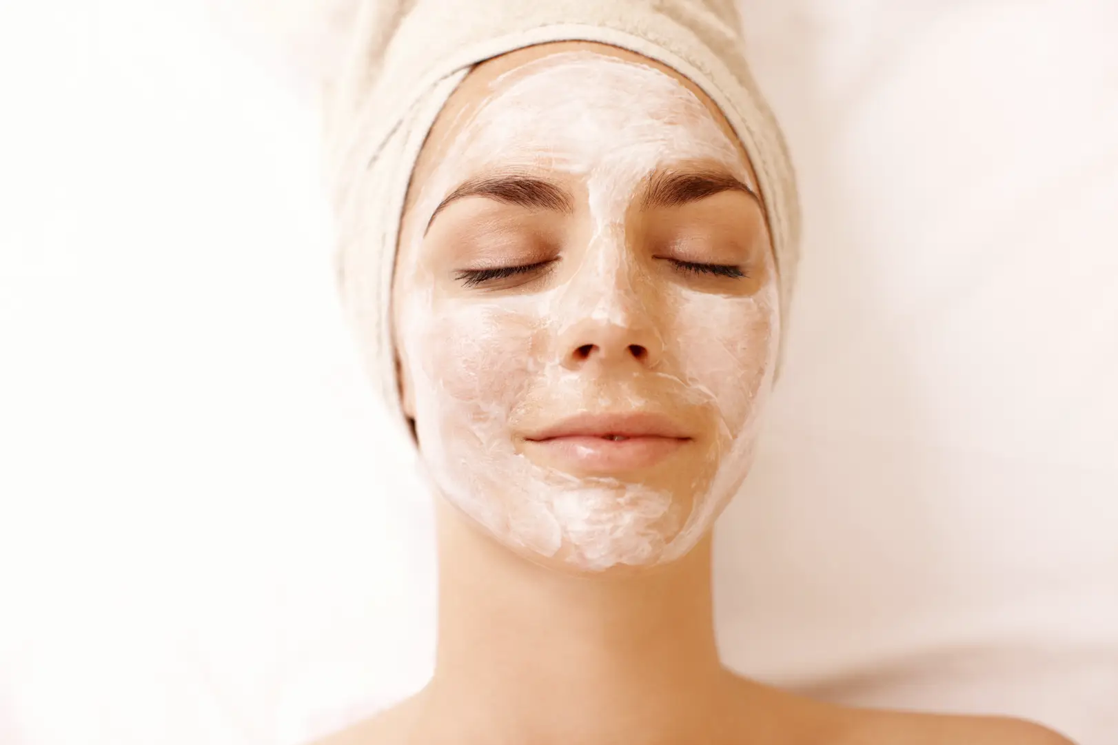 Woman with closed eyes wears a white facial mask and a towel wrapped around her hair.